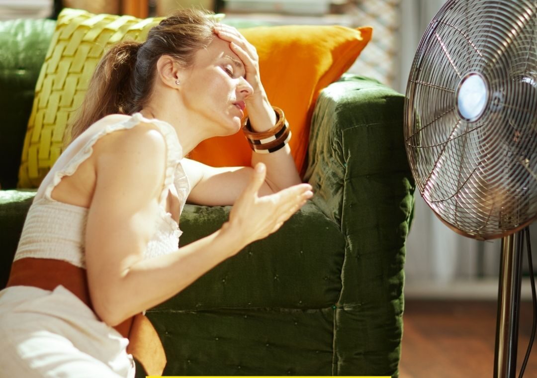 Une femme épuisée se tient devant un ventillateur, illustrant les difficultés physiques causées par la canicule