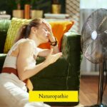 Une femme épuisée se tient devant un ventillateur, illustrant les difficultés physiques causées par la canicule
