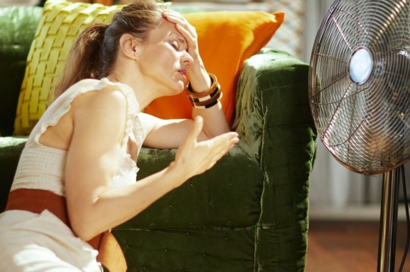 Une femme épuisée se tient devant un ventillateur, illustrant les difficultés physiques causées par la canicule